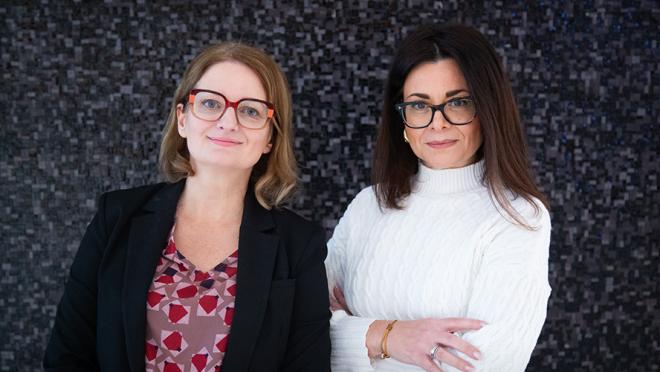Smiling women dressed professionally, standing in front of dark, blank wall.