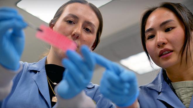 Kyla Bruce and Rachel Kuah in the lab wearing lab coats and looking at an experiment sample.