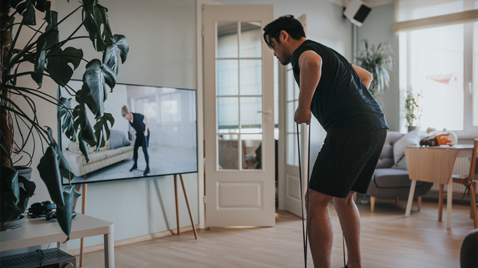 Male wearing fitness clothing performing an exercise with a resistance band in front of a TV screen in a living room. On the screen, a woman performs the same exercise beside a sofa.