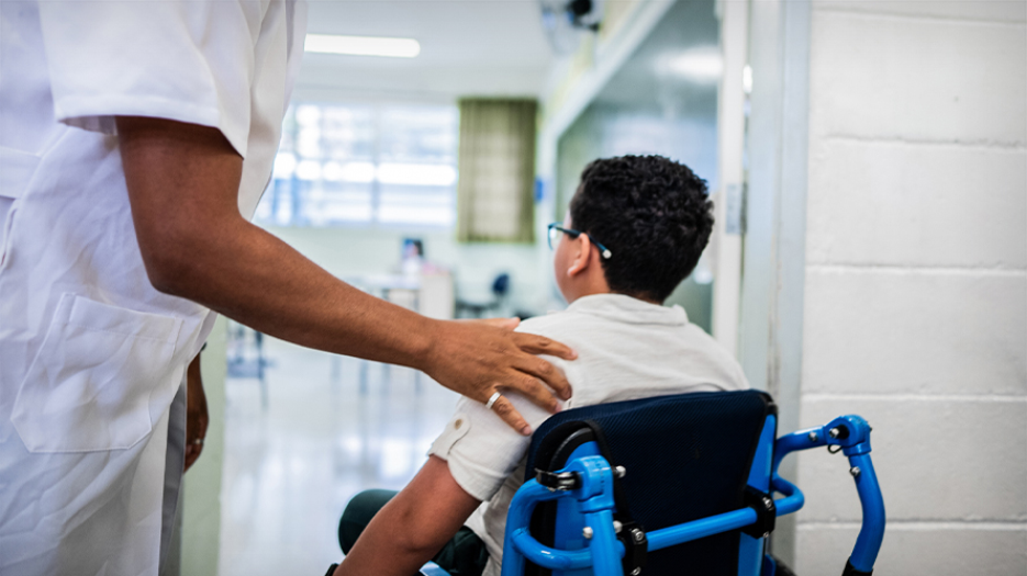 Child in wheelchair entering a classroom. An  adult, outside of the shot, places a hand on the child's shoulder as they enter.