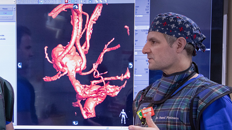 Male doctor in scrub cap and lead-lined garments in front of screen showing brain angiogram