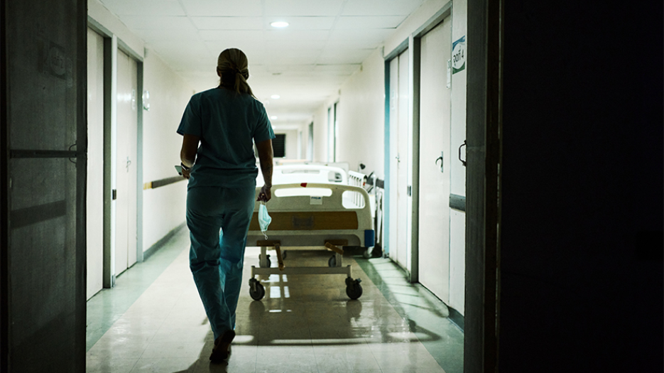 Female nurse in scrubs walking through a hospital corridor next to a hospital bed. The image is taken from behind the nurse, in a darkened hallway.