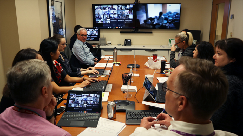 Group sitting around a conference room table participating in a video conference. Group attendees have computers in front of them and a large TV monitor can be seen at the front of the room. The image is taken from behind two individuals at the back of the room.