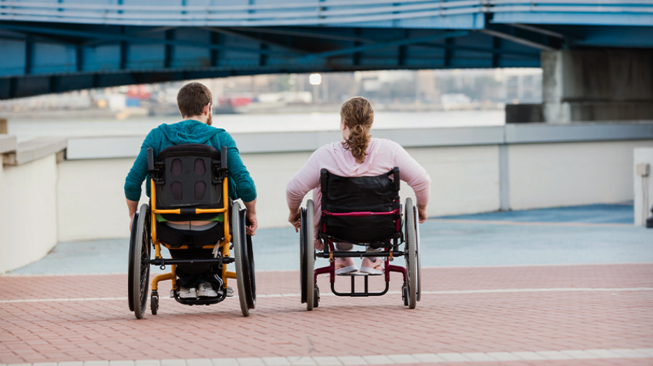 Two individuals using wheelchairs are seen from behind as they move along a paved walkway near the waterfront. One individual is male and wearing a teal hooded sweatshirt, the other is female and wearing a pink hooded sweatshirt.