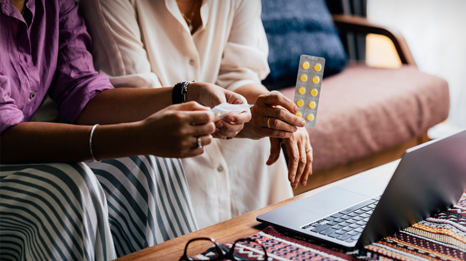 Close up shot of two individuals sitting on a couch and holding medication in front of an open laptop.