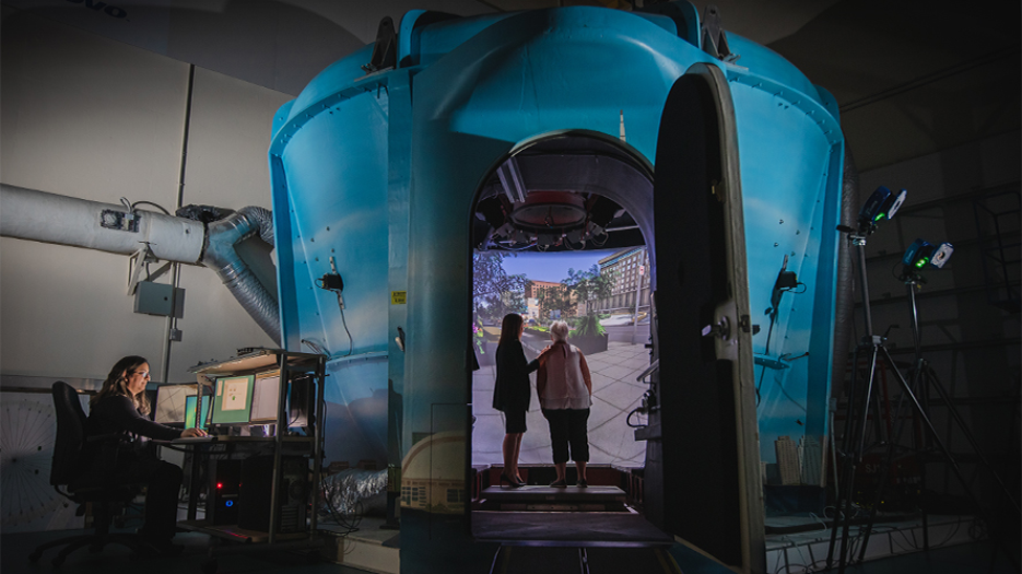 Wide angle shot of the KITE StreetLab, an immersive virtual reality facility,  with two females pictured inside as the screen displays a virtual street crossing.